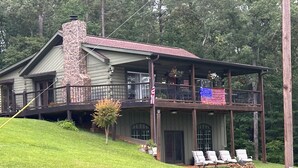 A two-story log cabin with a stone chimney, a balcony with a flag, and a lawn with lounge chairs.
