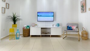 Ein modernes Wohnzimmer mit einem weißen Konsolentisch, einem Flachbildfernseher, einem Korbkistchen, einer Topfpflanze und einem dekorativen Sessel.