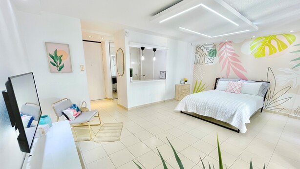 Ein modernes Schlafzimmer mit Bett, Nachttisch und Schreibtisch inklusive Computer. Ein Spiegel, eine Wand mit Blattmuster und eine Tür zu einem anderen Raum sind ebenfalls vorhanden.