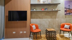 A modern room with a TV mounted on a wooden panel wall, two orange chairs with cushions, a small wooden table, and shelves with various items and a cityscape painting. - Condo Unit in Uptown Parksuites T2 Bgc (Taguig)