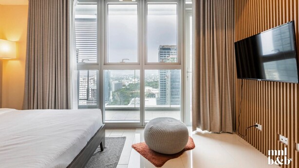 A modern hotel room with a large bed, a television, and a view of the city through floor-to-ceiling windows. - Condo Unit in Uptown Parksuites T2 Bgc (Taguig)