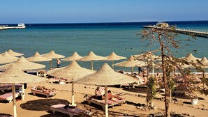 A beach with sun loungers and thatched umbrellas, a pier extending into the sea, and clear blue skies.