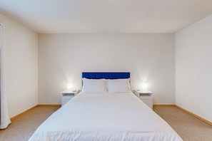 A neatly made bed with a blue headboard in a simple, clean room with white walls and a beige carpet.