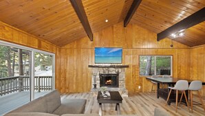 A cozy cabin interior with a stone fireplace, a large window overlooking a deck, and a dining area with chairs. - Crestline Lake Gregory Cabin  (Crestline)
