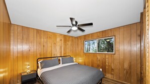 A bedroom with a bed, a nightstand, a ceiling fan, and a window with a view of trees. - Crestline Lake Gregory Cabin  (Crestline)