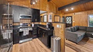 A modern kitchen with stainless steel appliances, wooden walls, and a gray sofa in a cozy living area. - Crestline Lake Gregory Cabin  (Crestline)