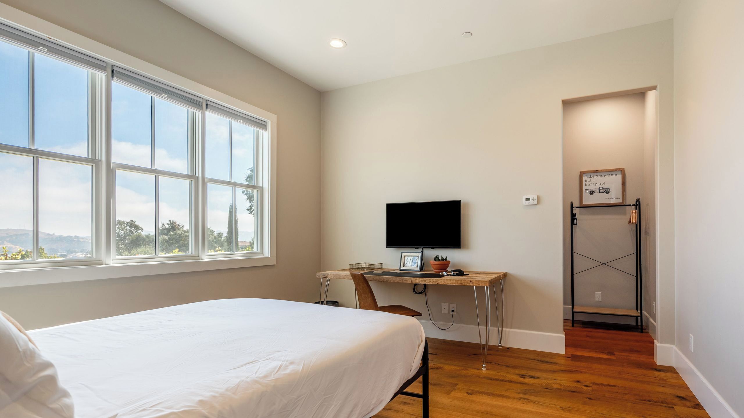 A bedroom with a bed, a desk with a computer, a TV, and a door leading to another room.