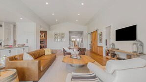 Uma sala de estar ampla com uma mesa de centro de madeira, sofás de couro, uma poltrona branca e uma cozinha com armários brancos.