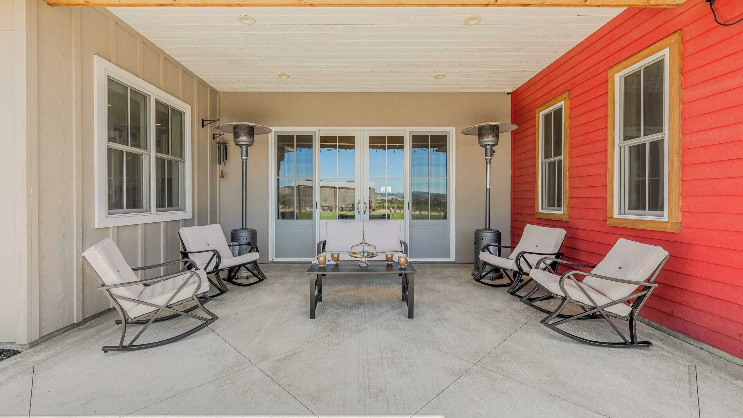 A covered patio with a table and four chairs, large windows, and a red wall.
