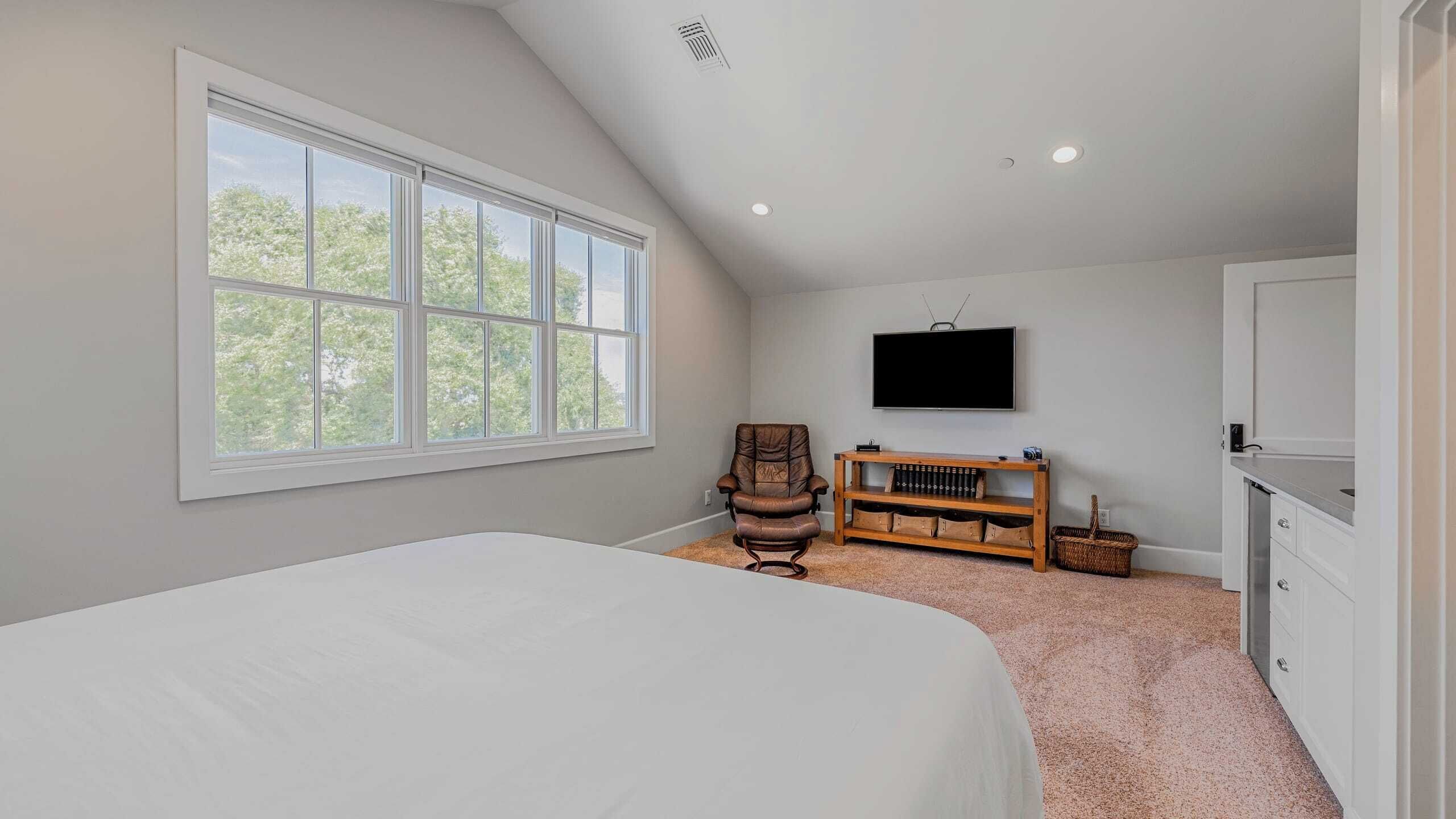 A bedroom with a large window, a bed, a chair, a TV, and a wooden shelf with books.