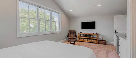 A bedroom with a large window, a bed, a chair, a TV, and a wooden shelf with books.
