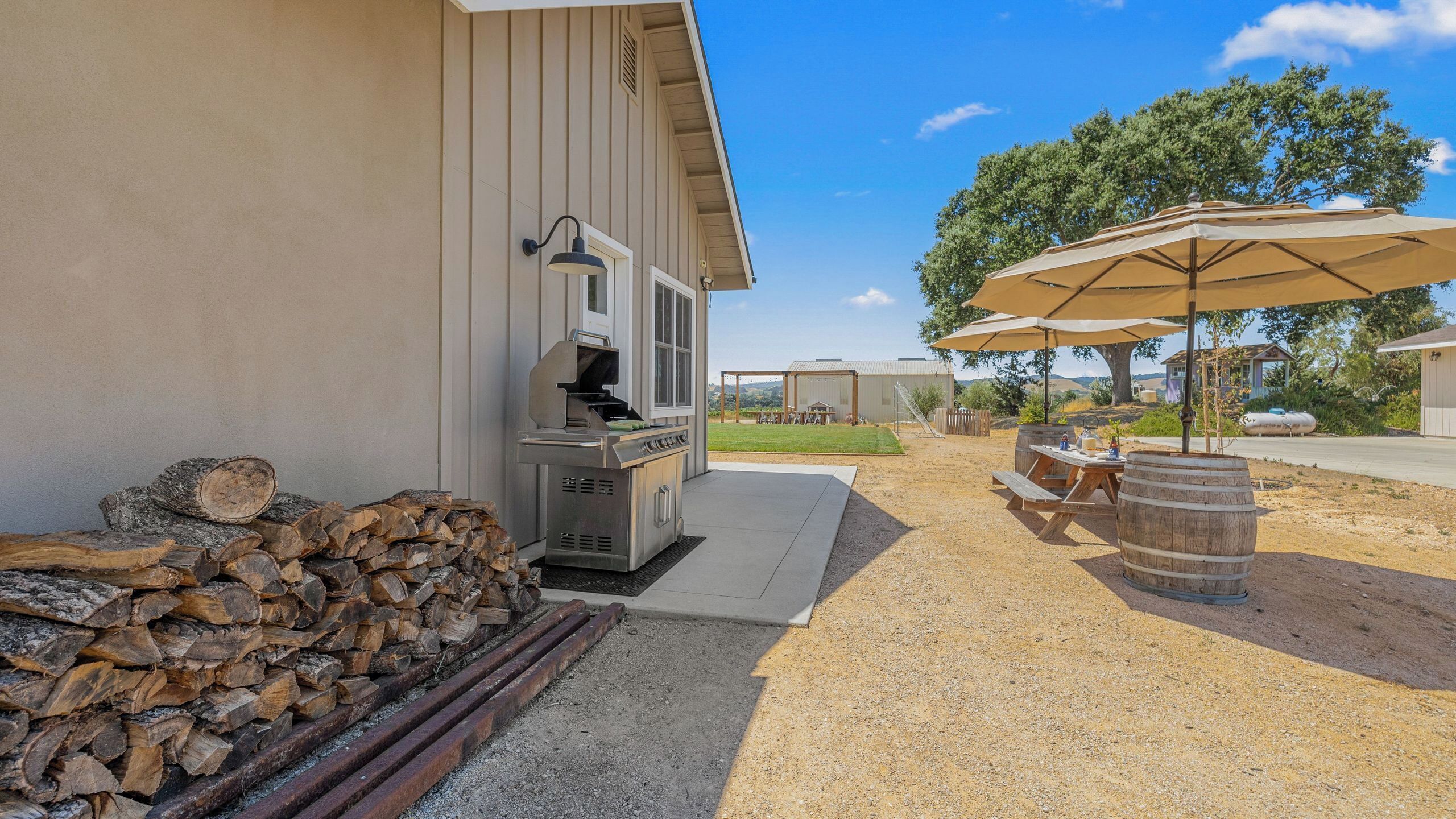 A barbecue grill, a stack of firewood, a picnic table with barrels, and an umbrella.