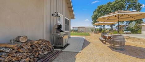 A barbecue grill, a stack of firewood, a picnic table with barrels, and an umbrella.