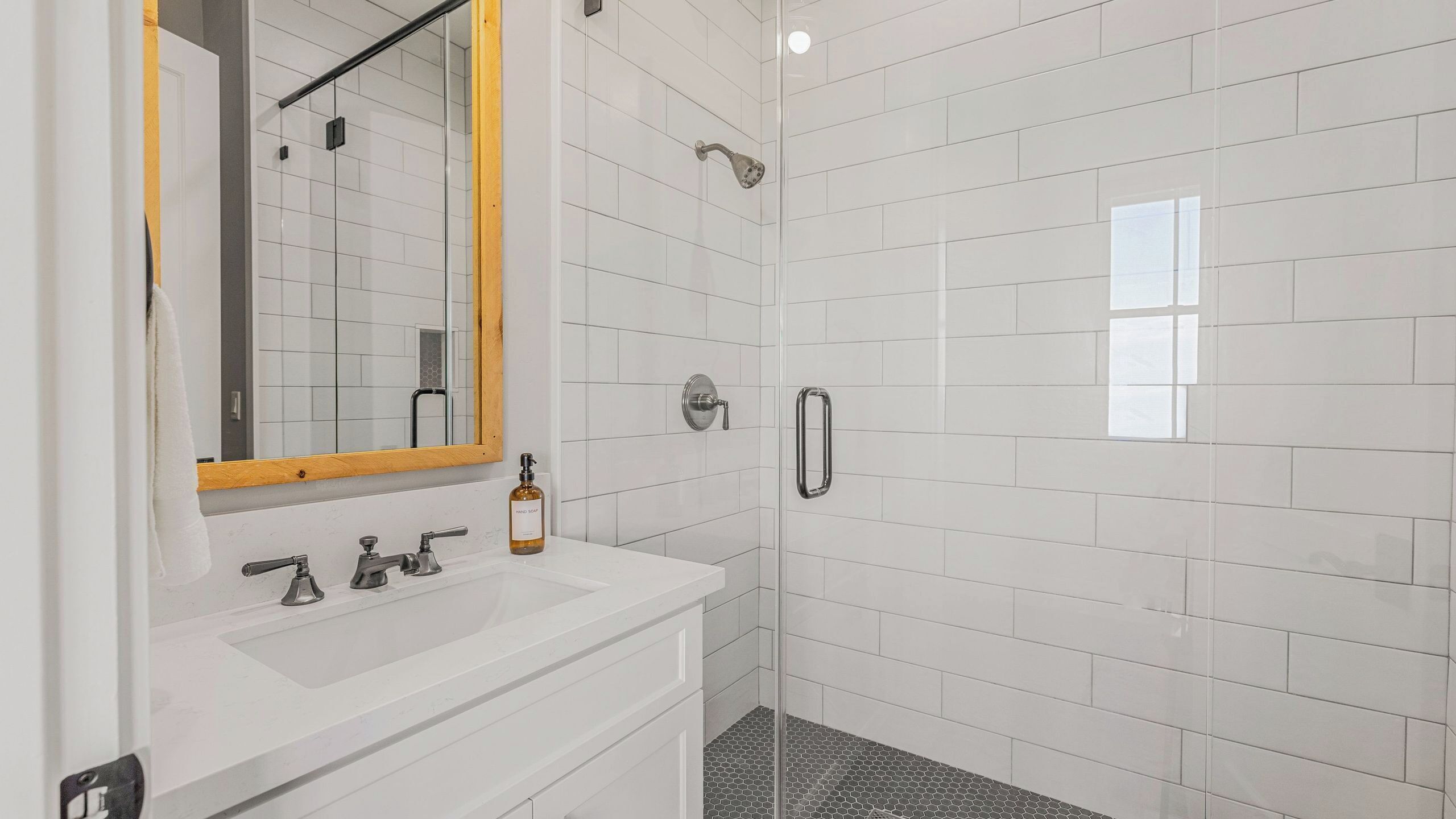 A bathroom with a white tiled shower, a sink with a mirror, and a bottle of hand soap.