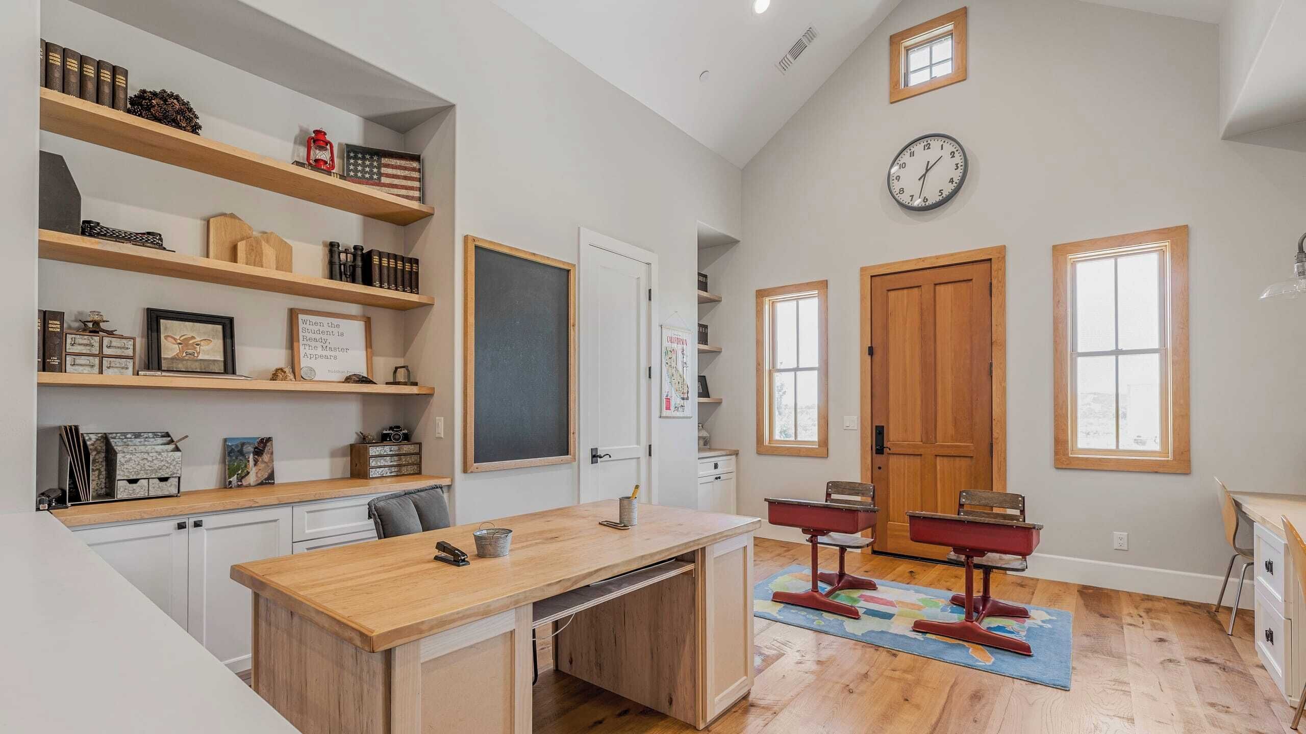 A spacious room with a wooden desk, shelves with books and decorative items, a chalkboard, and a large window.