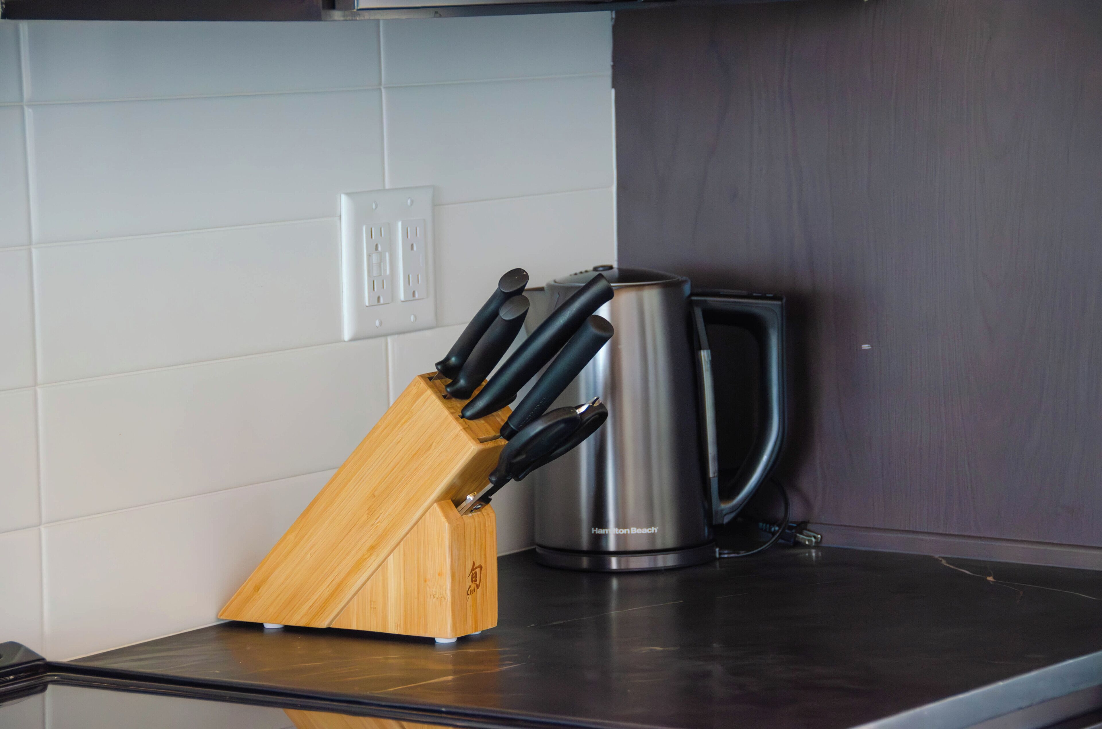 A kitchen counter with a knife block, a stainless steel kettle, and a wall socket.