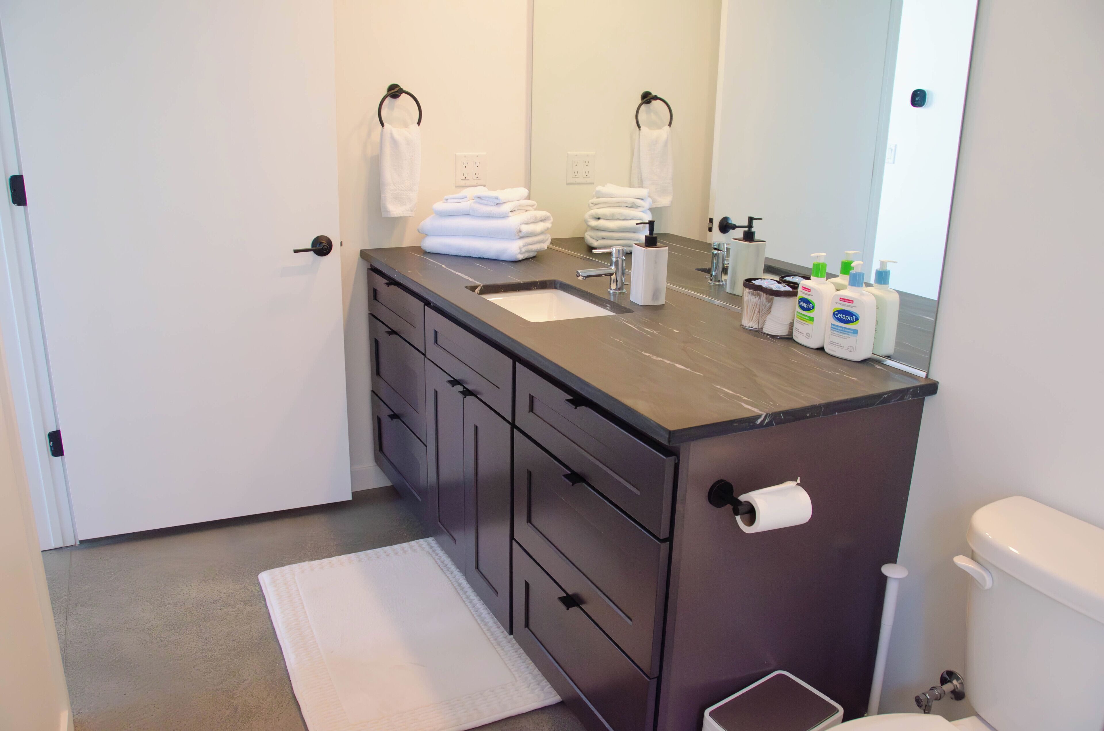 A modern bathroom with a dark countertop, a sink, a toilet, and a mirror. There are towels stacked on the countertop and toiletries on the sink.