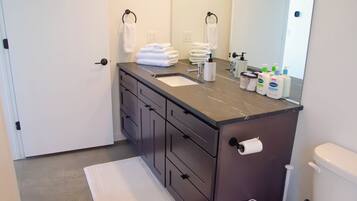 A modern bathroom with a dark countertop, a sink, a toilet, and a mirror. There are towels stacked on the countertop and toiletries on the sink.