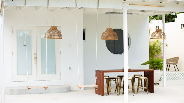A covered outdoor seating area with a wooden table and gold bar stools, surrounded by wicker pendant lights. - Top Location, 1 mile from Airport ,Full House, Big Patio, Free Parking. (Miami)