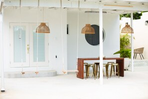 Un espace extérieur abrité, doté d’une table en bois et de tabourets de bar dorés, et entouré de suspensions en rotin.