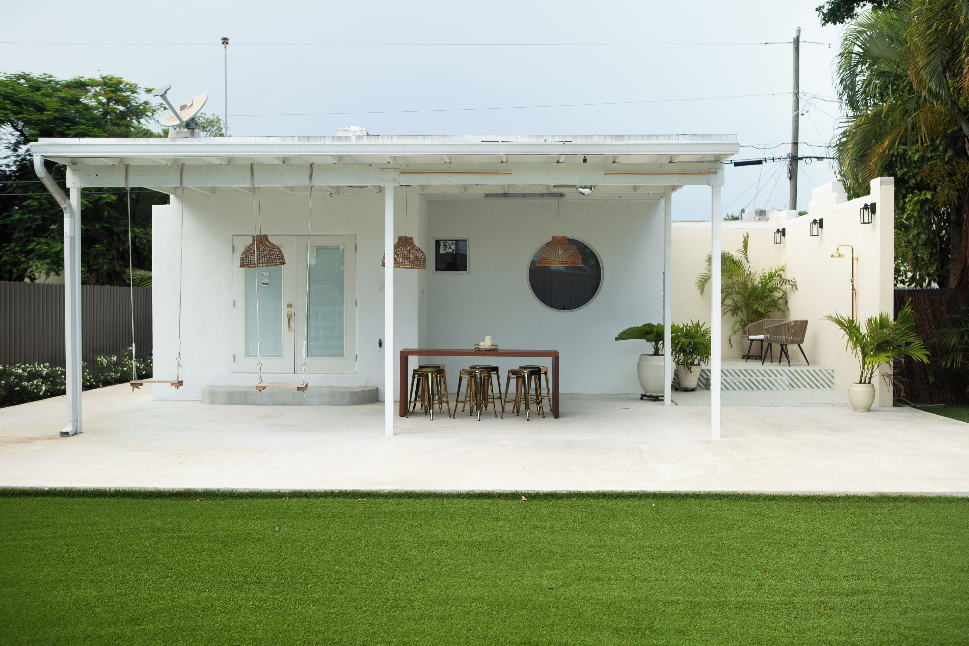 A covered outdoor patio with a table and stools, surrounded by potted plants and a white fence.