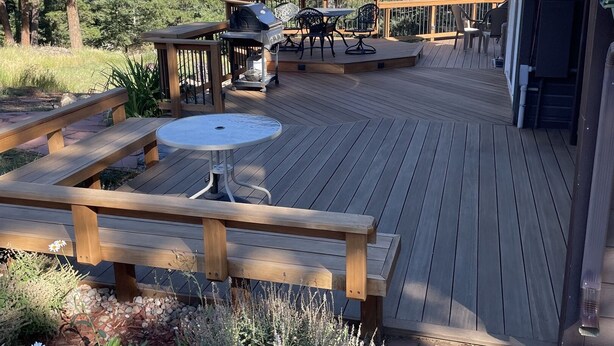 Eine Holzterrasse mit Grill, Tisch und Stühlen, umgeben von Grünpflanzen und Bäumen.