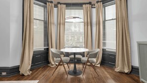 Une pièce d’angle avec une table ronde et quatre chaises, de grandes fenêtres équipées de stores et un luminaire au plafond.