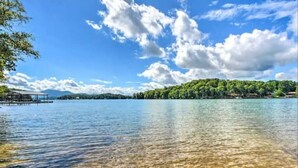A clear lake with a dock extending into the water, surrounded by lush greenery and a mountainous backdrop. - Tranquility at Claire de Lune Mountain Lake Home (Hayesville)