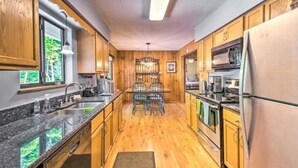 A kitchen with wooden cabinets, a granite countertop, a stainless steel refrigerator, and a dining area with a chandelier. - Tranquility at Claire de Lune Mountain Lake Home (Hayesville)