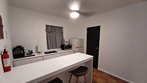 Uma cozinha compacta com bancada branca, micro-ondas, cafeteira e frigorífico preto. Há dois bancos e um extintor de incêndio na bancada.