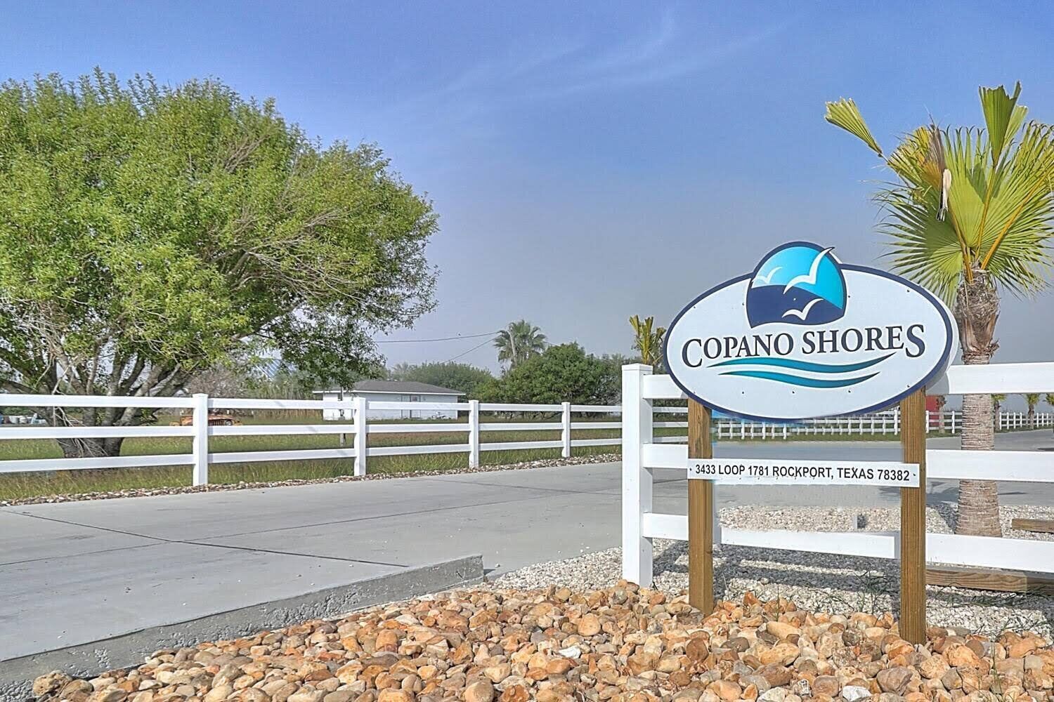 A sign for 'Copano Shores' with a logo, address, and a view of a gated area with trees and a fence.