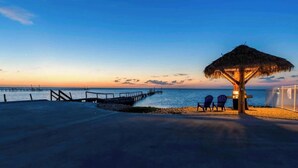 A beach scene with a thatched-roof structure, two chairs, and a pier extending into the water. - Paradise on Copano Bay ~ Complementary Kayaks ~ Pier and Pool  (Rockport)