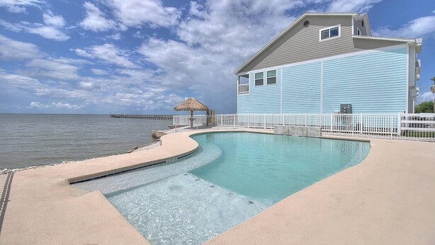 A pool area with a curved edge, a thatched-roof structure, and a building with a light blue exterior. - Paradise on Copano Bay ~ Complementary Kayaks ~ Pier and Pool  (Rockport)