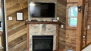 A cozy living room with a stone fireplace, a flat-screen TV mounted above it, and a wooden dining table. - Owls Nest Silo - 100 y/o converted concrete silo (Elmdale)