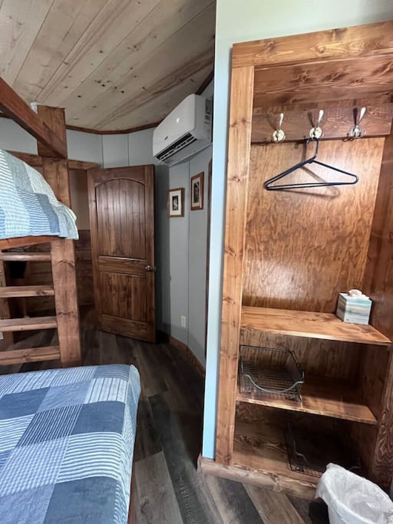 Un dormitorio pequeño con literas, un armario de madera y un estante de madera con un perchero y una caja de pañuelos.