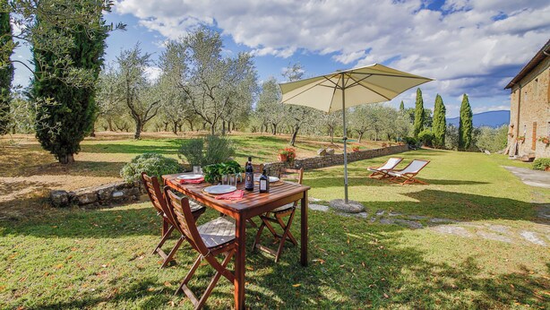 Un espace extérieur aménagé pour les repas, avec une table et des chaises, un grand parasol et une vue sur un jardin agrémenté d’arbres et d’un mur en pierre.
