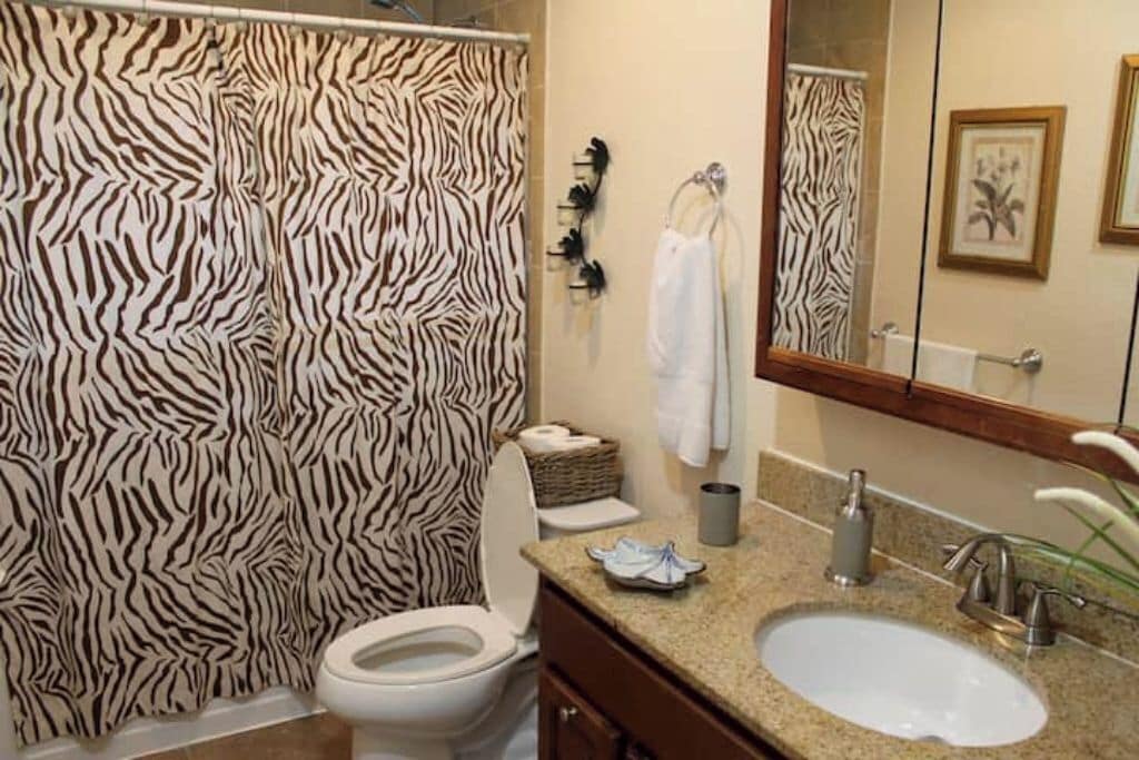 Um banheiro com cortina de chuveiro listrada como zebra, vaso sanitário branco, pia com espelho e porta-toalhas.