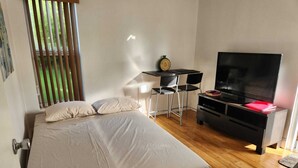 A bedroom with a bed, a desk, a chair, a television, and a window with blinds.