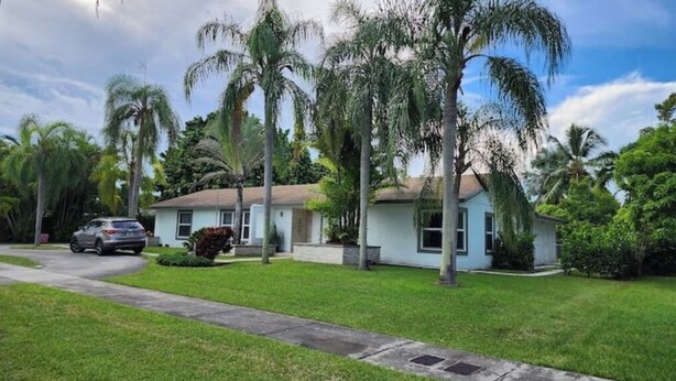 A single-story house with a light blue exterior, surrounded by lush greenery and palm trees.