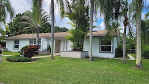 A single-story house with a white exterior and a brown roof, surrounded by a well-maintained lawn and several palm trees.