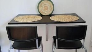 A small dining area with two black chairs, a table with a woven placemat, and a decorative vase with green foliage.