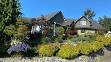 A two-story house with a stone retaining wall, surrounded by various shrubs and trees.