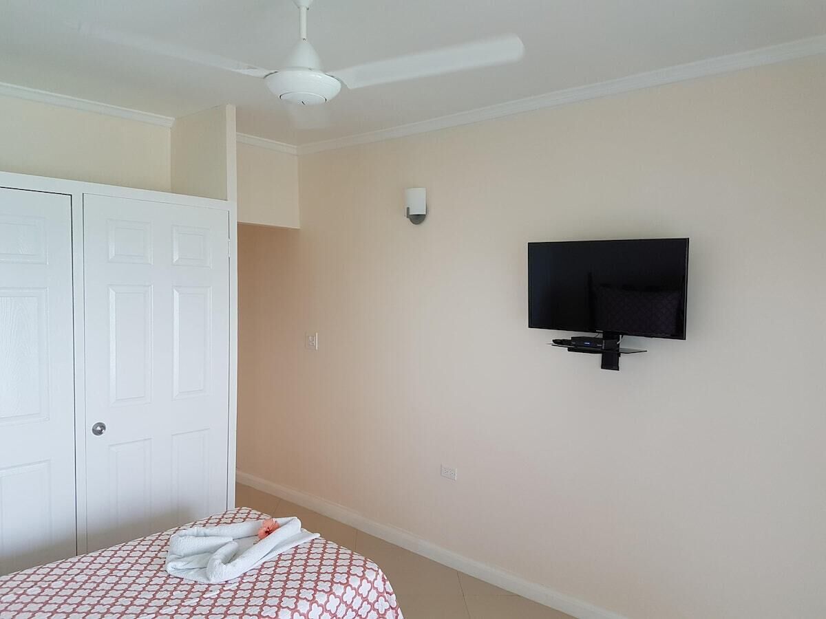 A bedroom with a bed, a white wardrobe, a ceiling fan, a wall-mounted TV, and a light switch.