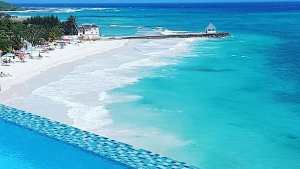 A beach with a clear blue ocean, a white sandy shore, and a small building near the water. - The Happy Place- 4BR Beachfront Fully Staffed Villa w/ Eternity Pool (Montego Bay)