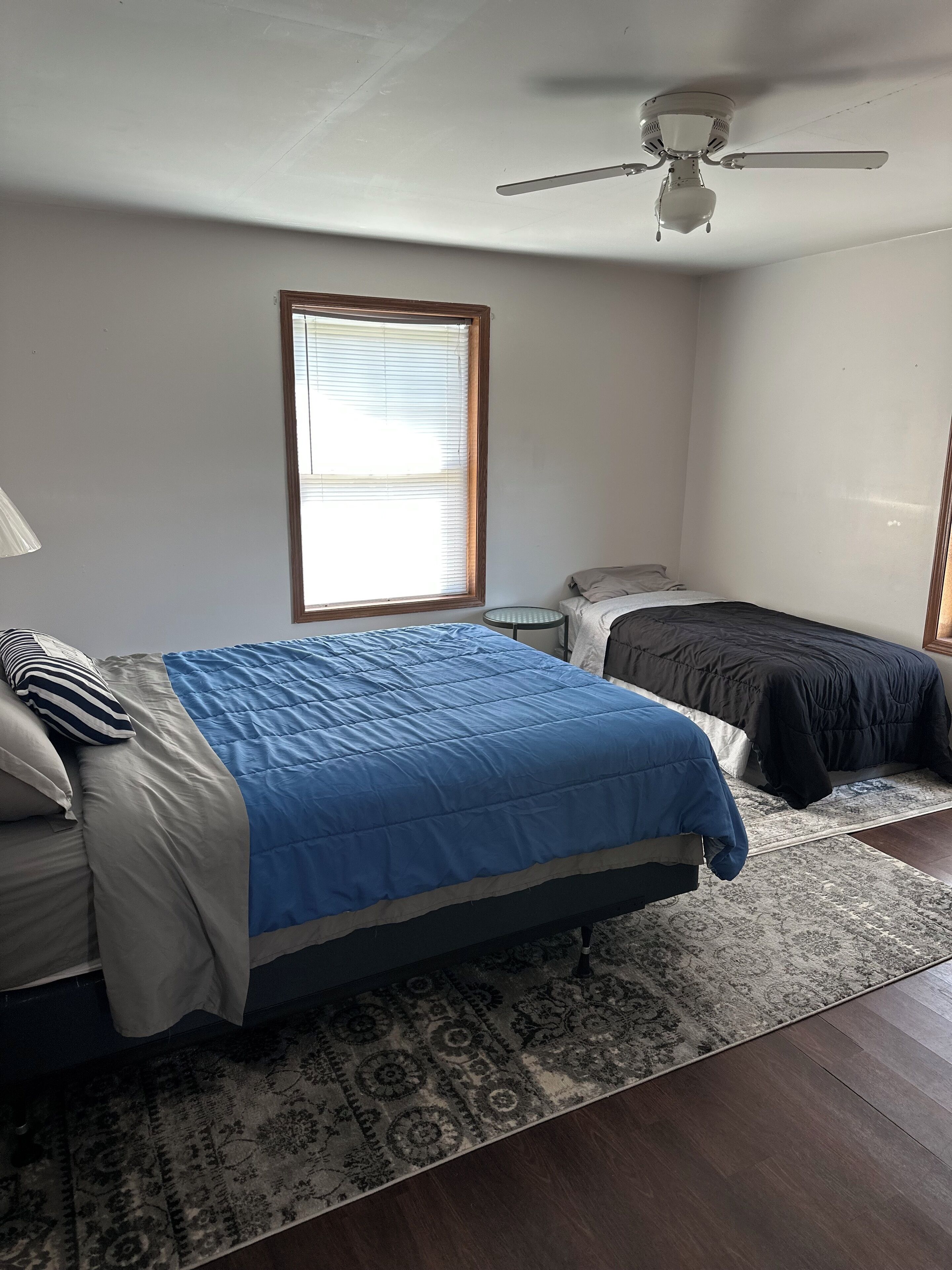 Une chambre avec deux lits, un ventilateur de plafond, une fenêtre avec des stores et un tapis à motifs.