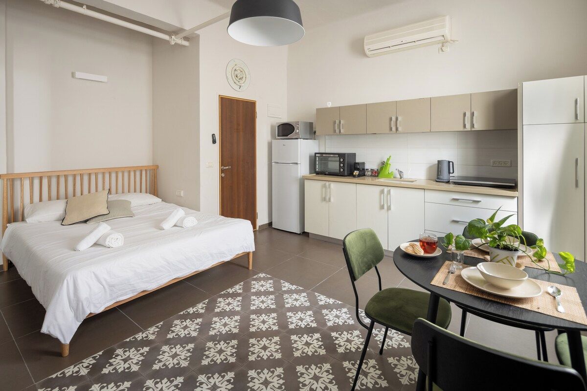 A bedroom with a bed, a dining table with chairs, a kitchen area with appliances, and a small potted plant.