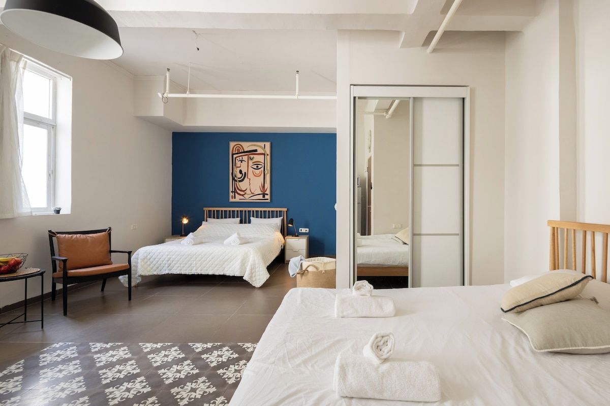 Une chambre d’hôtel avec un grand lit, deux petits lits, une chaise, un bureau et un mur orné d’un tableau.