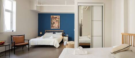A hotel room with a large bed, two smaller beds, a chair, a desk, and a wall with a painting.