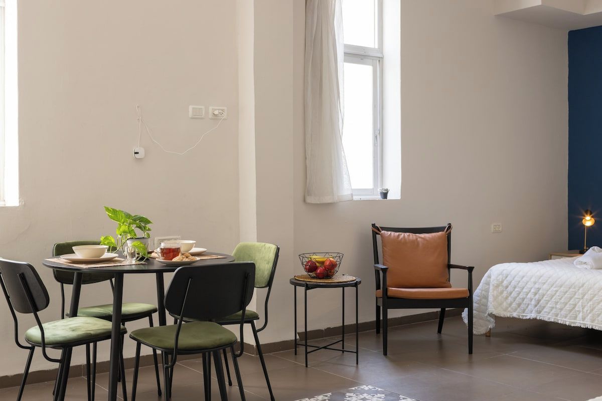 A room with a dining table, chairs, a small side table with a bowl of fruit, a bed with a white quilt, and a window with a curtain.
