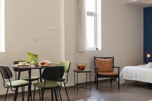 Une pièce comprenant une table à manger, des chaises, une petite table d’appoint avec un saladier de fruits, un lit recouvert d’une courtepointe blanche et une fenêtre avec un rideau.